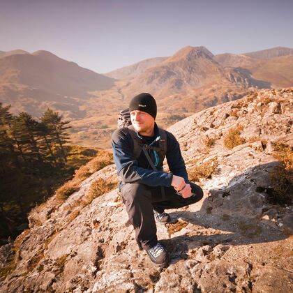 A man in winter walking gear crouching on a rocky outcrop in the Welsh mountains.