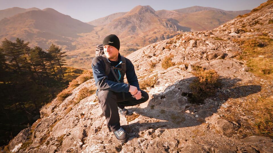 A man in winter walking gear crouching on a rocky outcrop in the Welsh mountains.