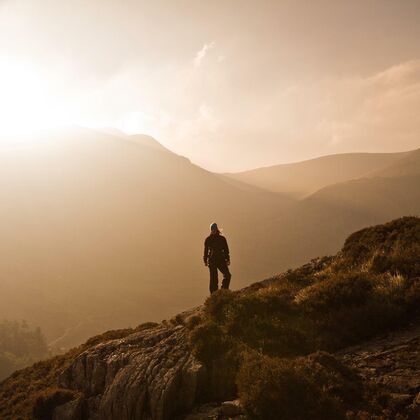 A mountainous landscape vista with the setting sun silhouetting a lone hiker on a ridge.