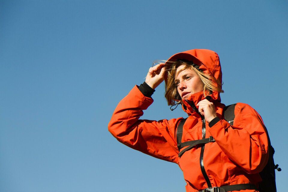A young woman in red hiking jacket raises her hood, no other details can be seen except a clear blue sky.