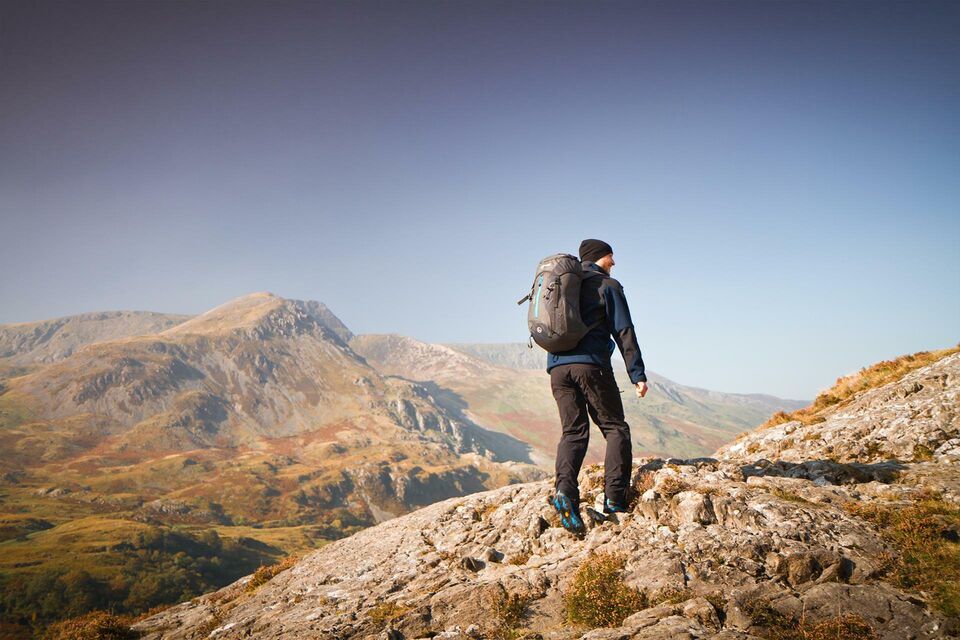 A lone make hiker with backpack walks away from the camera and toward the Welsh mountains.