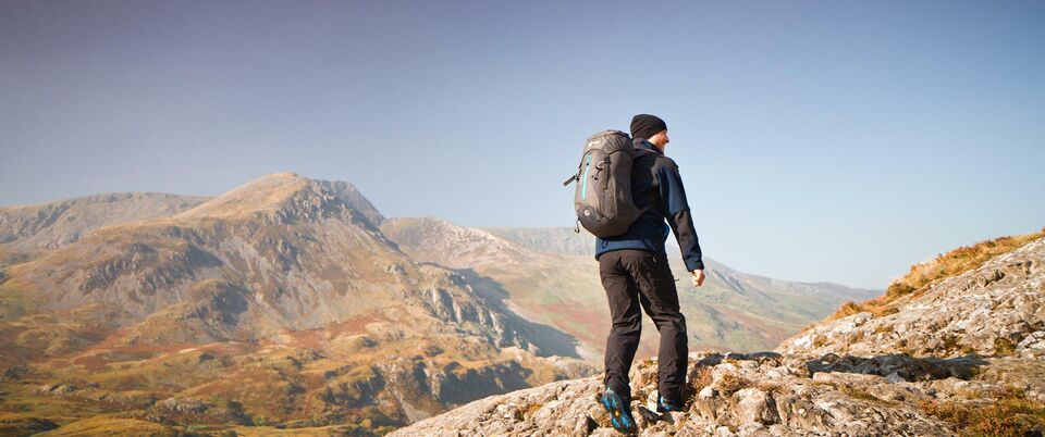 A lone make hiker with backpack walks away from the camera and toward the Welsh mountains.